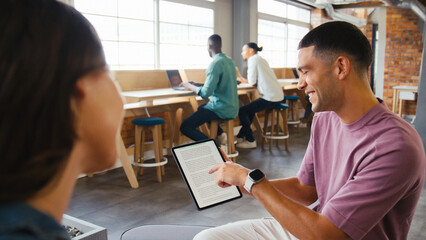 Two Young Businesspeople With Digital Tablet Meeting In Office With Colleagues In Background