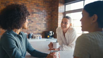 Multi-Cultural Businesswomen Taking Coffee Break In Kitchen Area Of Modern Open Plan Office Together