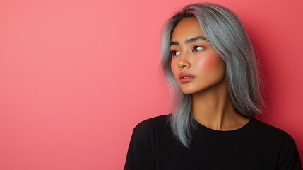 Fototapeta premium A young Asian woman with silver-blue hair in long layers, wearing a black t-shirt, gazes thoughtfully to the side against a pastel pink background