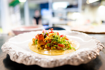A gourmet dish elegantly presented on a white ornate plate, with a blurred restaurant kitchen in the background, highlighting the culinary art and sophisticated dining ambiance.