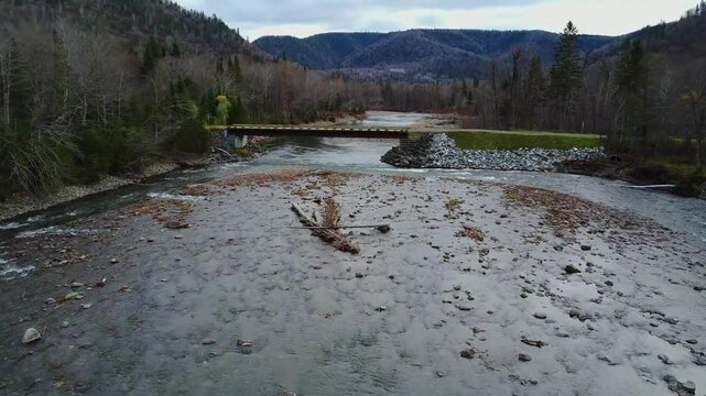 Un drone survol le lit rocheux d'une rivi&egrave;re sauvage parsem&eacute; d'arbres morts qui passe sous un pont forestier dans un paysage de for&ecirc;t automnale montagneuse. ( Rivi&egrave;re Anger, Comt&eacute; de Bonaventure Qc )
