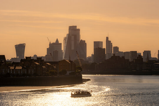 Fototapeta A riverboat peacefully sails on a river during sunset, with a backdrop of a modern city skyline, featuring diverse architectural styles and high-rise buildings in London
