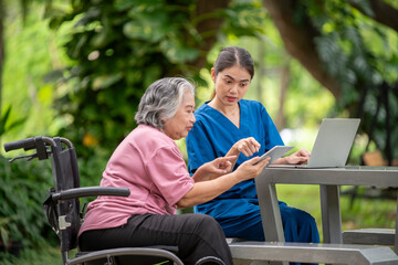 Elder woman is sitting a wheelchair holding a tablet, while a young healthcare professional standing smiling look tablet likely explaining something .They are outdoor park healthy treatment concept.