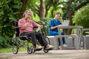 Elder woman is sitting a wheelchair holding a tablet, while a young healthcare professional standing smiling look tablet likely explaining something .They are outdoor park healthy treatment concept.
