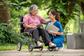 Elder woman is sitting a wheelchair holding a tablet, while a young healthcare professional standing smiling look tablet likely explaining something .They are outdoor park healthy treatment concept.