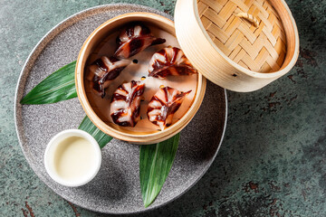 A ceramic plate with a bamboo steamer, green leaves, and a small bowl of sauce on a textured surface. Ideal for Asian cuisine, food presentation, and culinary themes. High-quality, natural light.