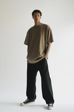 Young Man in Oversized Brown T-Shirt and Black Pants Posing Against White Background