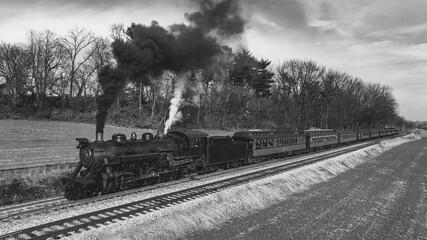 A vintage steam locomotive chugs along the tracks, releasing thick black smoke into the sky while passing through a serene countryside dotted with trees and open fields.