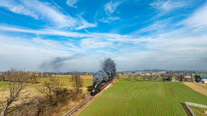 A steam locomotive chugs along railway tracks, billowing dark smoke against a backdrop of lush fields and a clear blue sky. The serene landscape features open fields and distant hills.
