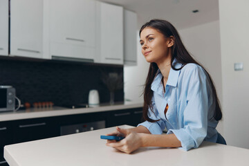 Woman sitting at table in kitchen with a cell phone in hand, looking thoughtful and relaxed