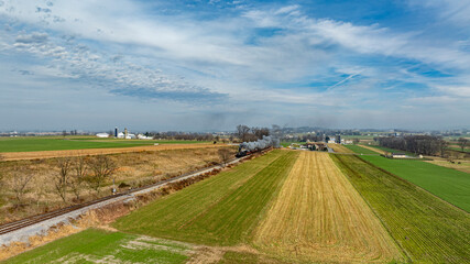 A steam train chugs along railway tracks, passing vibrant green and golden fields. Farm buildings and a distant landscape add to the scenic rural charm of this tranquil countryside setting.