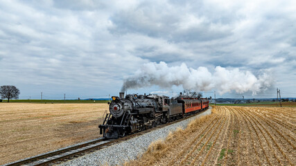 Naklejka premium A vintage steam train chugs along railway tracks, releasing plumes of vapor as it passes through golden fields of harvested crops under a gray, overcast sky.