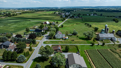 This view showcases a serene rural community with houses and farms nestled among lush green fields under a clear blue sky, embodying the tranquility of summertime.