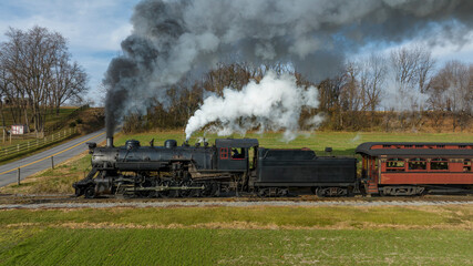 A classic steam locomotive is moving along tracks surrounded by lush green fields and trees, billowing smoke into the clear blue sky on a beautiful day.