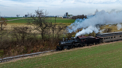 A vintage steam locomotive pulls a passenger train along a scenic rural track, surrounded by fields and distant barns. The sky is overcast, giving a moody ambiance to the picturesque landscape.