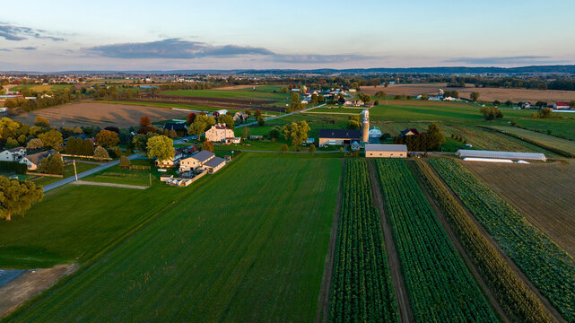 The view captures a peaceful rural community with expansive fields, scattered homes, and a silvery sunset. The rich greenery and orderly agricultural layout highlight the area serenity.