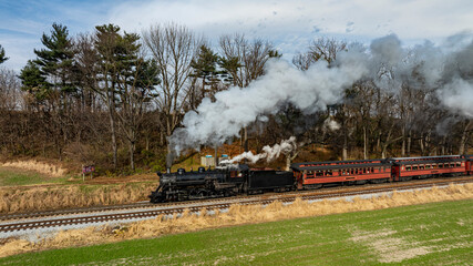 A classic steam locomotive billows smoke as it travels along the railway, framed by greenery and clear skies, showcasing the beauty of a bygone era in transportation.