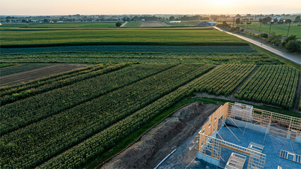 An expansive view showcases lush agricultural fields with varying crop patterns along with a construction site. The warm hues of the sunset illuminate the serene rural landscape.