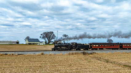 A classic steam locomotive chugs along the tracks, trailing puffs of smoke as it passes by a quaint farmhouse and fields under a cloudy sky