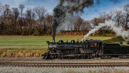 A steam locomotive emits clouds of smoke as it travels along railway tracks in a scenic countryside. The vibrant natural surroundings highlight the beauty of autumn foliage.