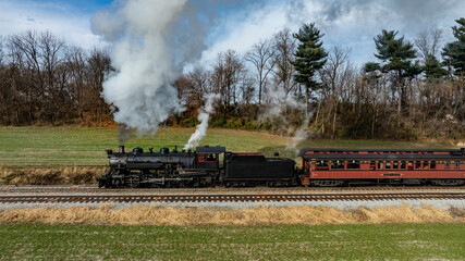 A classic steam locomotive chugs along the tracks, releasing puffs of white smoke into the clear blue sky. The vintage passenger car follows closely, representing a nostalgic journey.