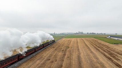 Obraz premium A vintage steam train chugs along tracks cutting through golden fields under a gray sky, with farmland visible in the distance, showcasing a serene rural landscape.
