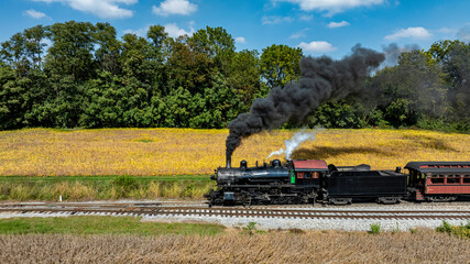 A steam locomotive emits black smoke as it moves along the tracks near a golden field and trees under a clear blue sky.