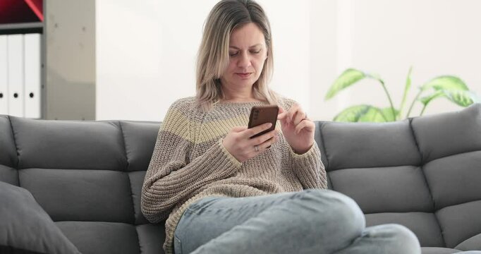Young woman scrolls through social media via phone sitting on sofa at home. Lady enjoying downtime in comfortable living space during weekends in apartments