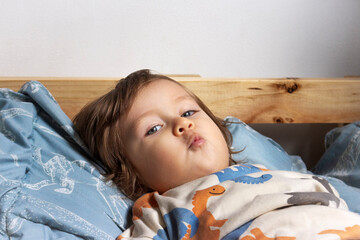 Niño rubio de dos años echado en su cama azul, él está mirando a la cámara, está haciendo una...