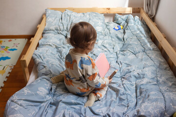 Bebé de un año sentado en su cama, él está pintando con sus colores sobre un cuaderno con hojas...