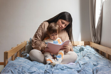 Mamá con bebé de un año sentado en sus piernas, ella le está leyendo un cuento, ellos están...