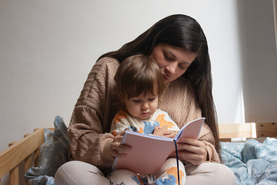 Mam&aacute; con beb&eacute; de un a&ntilde;o sentado en sus piernas, ella le est&aacute; leyendo un cuento, ellos est&aacute;n sentados sobre una cama. Actividad madre e hijo