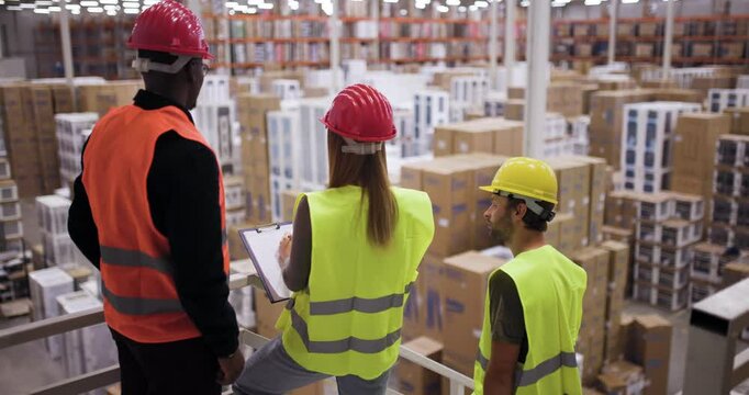 Warehouse workers observing stock organization while wearing safety gear including helmets and vests in a large distribution center full of shelves and boxes.
