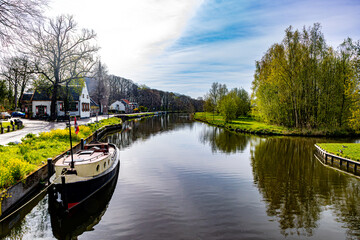Fototapeta premium Boat anchored on canal bank between houses and bare trees against blue sky in background, rural vehicular road along river, sunny spring day in Oud-Zuilen, Utrecht, Netherlands