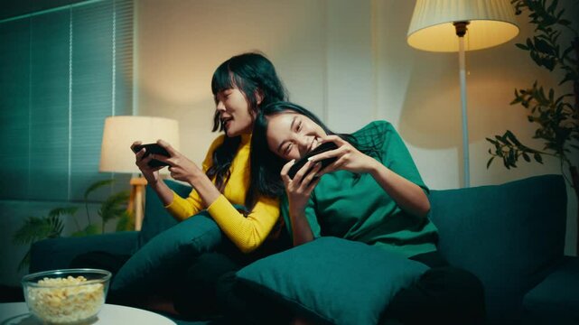 Two women are sitting on a couch in their living room, playing video games. They are both smiling and laughing and seem to be enjoying themselves