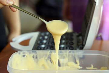 Hand holding a spoon, drop water and waffle batter onto the circle mold of a small electric waffle maker. Easy to make at home.