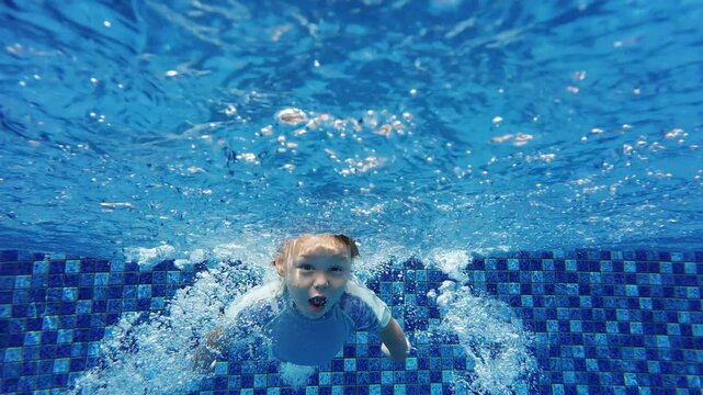 Fearless toddler dives into swimming pool, captured in slow motion. Young child, with open eyes and joyful expression, demonstrates boldness and determination as he paddles through water.