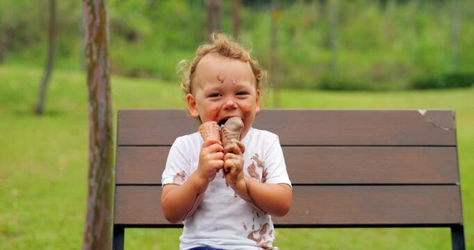 Cheerful young boy, seated on park bench, bursts out laughing as he indulges in double treat of messy ice cream. His face and clothes are smeared and stained with melting sweetness.