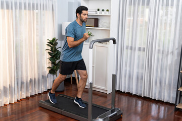 Fototapeta premium Athletic and sporty man running on running machine during home body workout exercise session for fit physique and healthy sport lifestyle at home. Gaiety home exercise workout training.