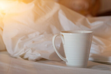 A white ceramic coffee mug was placed on the bed in the bedroom in the morning after the cup had been consumed. A white ceramic mug for hot tea and coffee is placed on a white cloth in the bedroom.