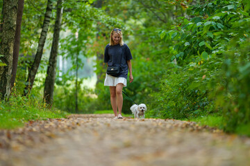 Fototapeta premium Girl walking the dog in the park forest. Rest concept. Woman walking with a dog on a leash in blue dress and with shoes.