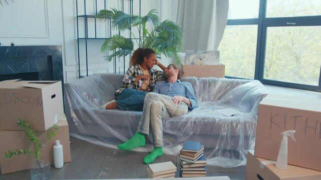 In a sunlit room, a diverse couple shares laughter on a couch surrounded by unpacked cardboard boxes. Their playful banter creates a warm atmosphere, filled with joy and anticipation.