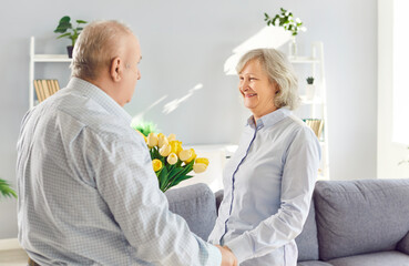 Happy cheerful senior woman getting tulips flowers bouquet from her husband. retired man making romantic present to his wife on marriage anniversary holiday standing in living room at home.