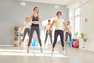 Group of happy cheerful beautiful sporty young women in sports clothes doing jumping exercises all together during an active fitness workout at the gym