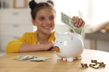 Pocket money. Cute girl putting dollar banknote into piggy bank at wooden table indoors, selective...