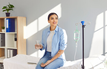 Beautiful young woman with sterile needle in arm sitting on medical bed by IV drip stand at clinic,...