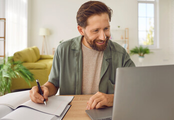 Happy adult man studying online. Smiling bearded male student in casual shirt sitting at desk at...