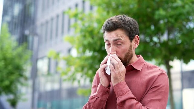 Man experiencing allergy symptoms sneezes into tissue while standing outside in urban environment. Businessman wears casual red shirt, surrounded by city buildings, trees. Allergy reaction during day