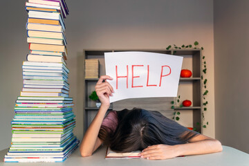 Overwhelmed student with head down holding a help sign. A student rests their head on a desk,...