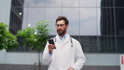 Smiling bearded doctor using phone while walking outside in front of a hospital building. Positive medical worker physician wears white coat, glasses, stethoscope. Represents healthcare and technology - Powered by Adobe
